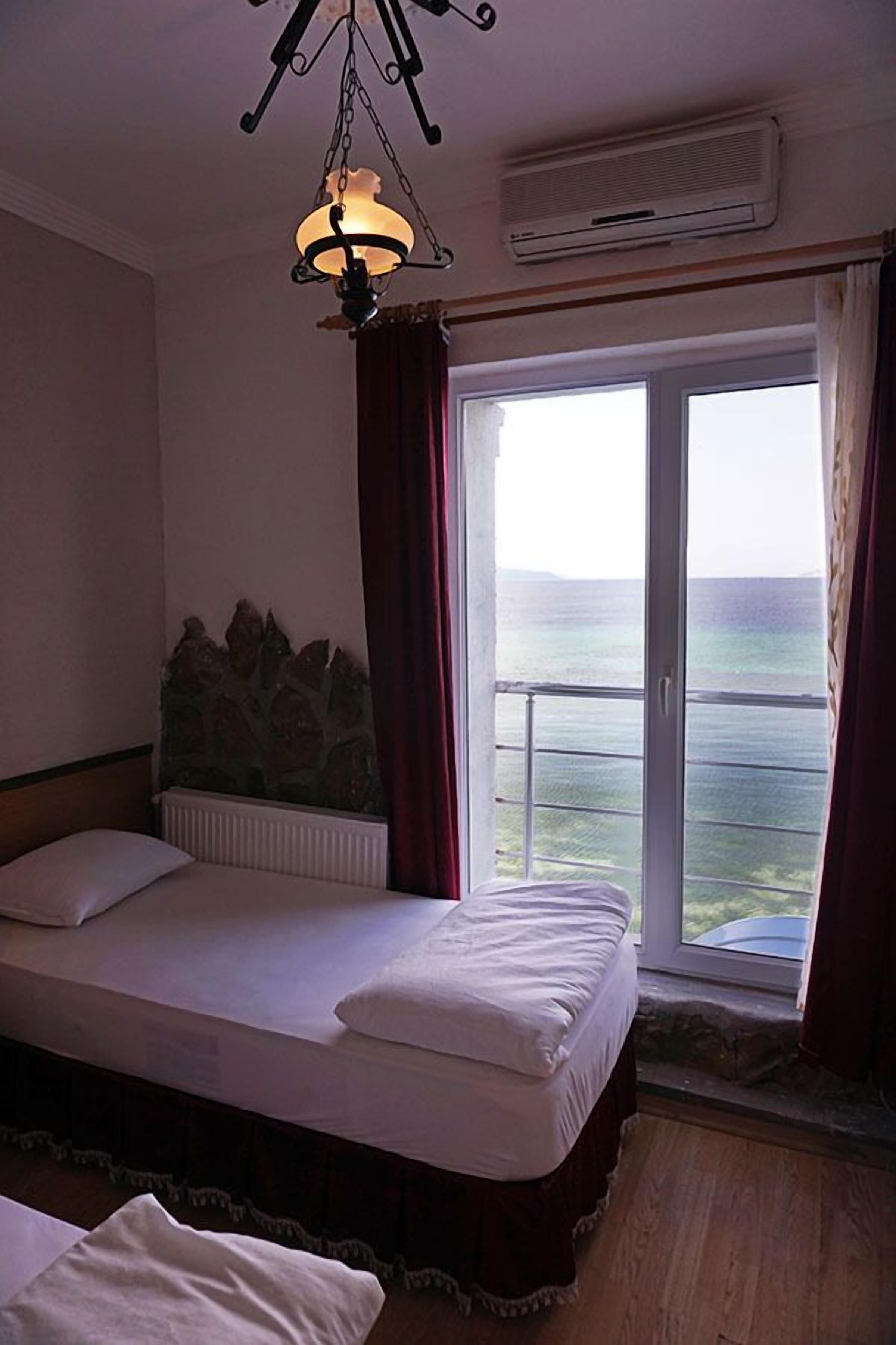 A bedroom with a bed, a stone headboard, a window with curtains, and a view of the sea.