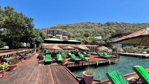 A wooden deck with green lounge chairs and umbrellas, a pool, and a building with a green roof in the background. - Veranda Otel Bozburun (+16) (Marmaris)