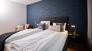 Ein ordentlich bezogenes Bett mit weißen Leinen und mehreren Kissen, ein Nachttisch mit einer Lampe und eine Wand mit geometrischen Mustern.