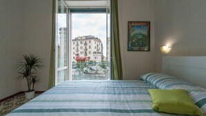 A hotel room with a bed, a potted plant, a painting, and a view of buildings through a window. - B&B Antonello (Salerno)