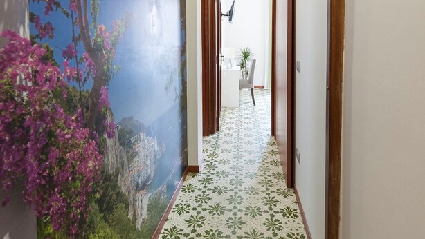 Un couloir avec des carreaux de sol à motifs, une fresque représentant des fleurs et de la verdure, et une vue sur une pièce où l’on aperçoit une chaise et une table.
