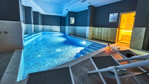 Una piscina interior con suelo de mosaico azul, rodeada de paredes y techo con iluminación empotrada.