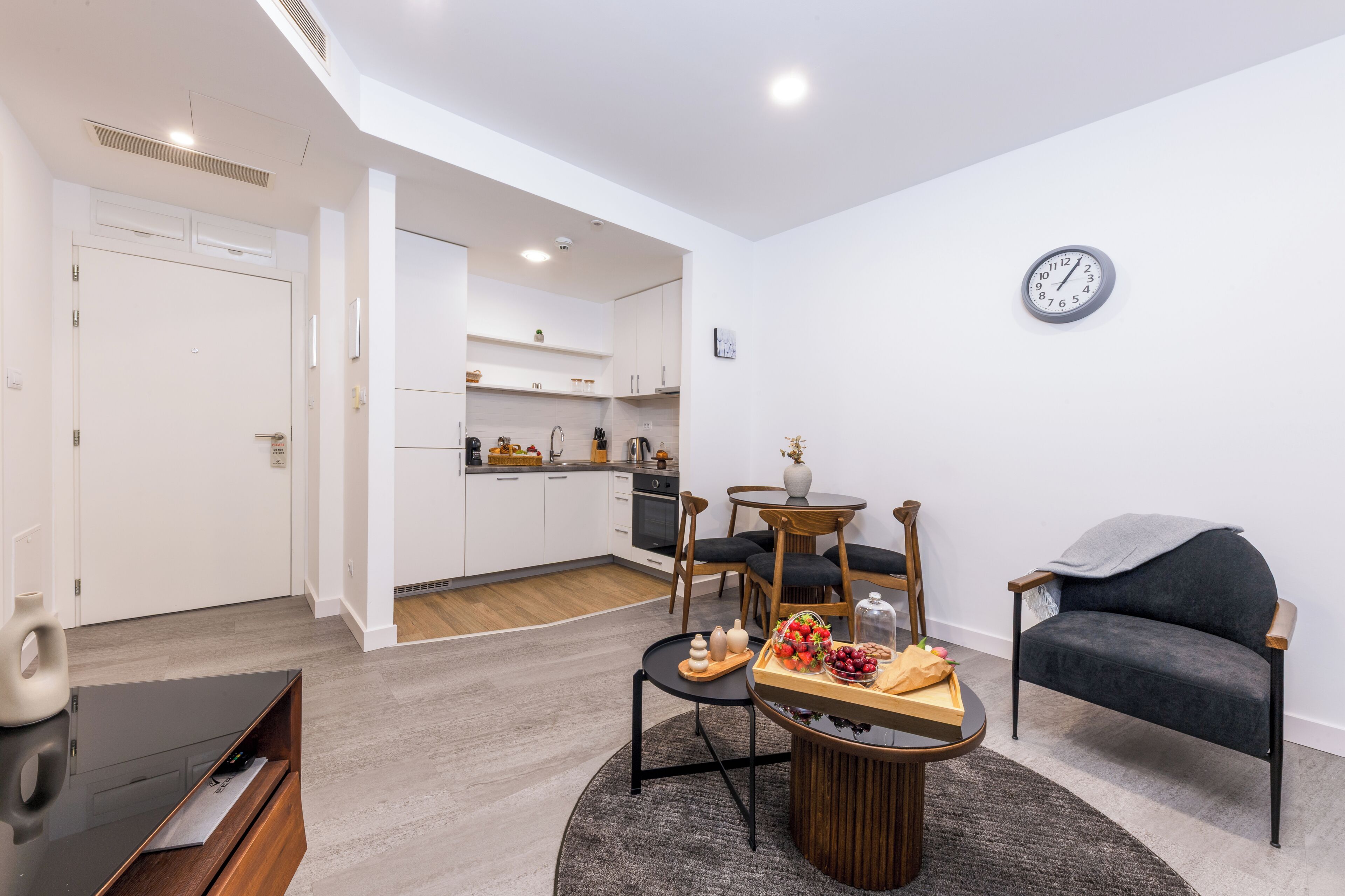 A modern living room with a TV, a dining table with a fruit tray, a sofa, and a kitchen in the background.