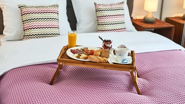 Una cama bien hecha con una manta rosa con textura, una bandeja de madera con artículos para el desayuno y una lámpara de noche.