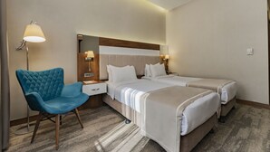 Una habitación de hotel con dos camas, un sillón azul, una mesita de noche con lámpara y un espejo grande sobre la cama.