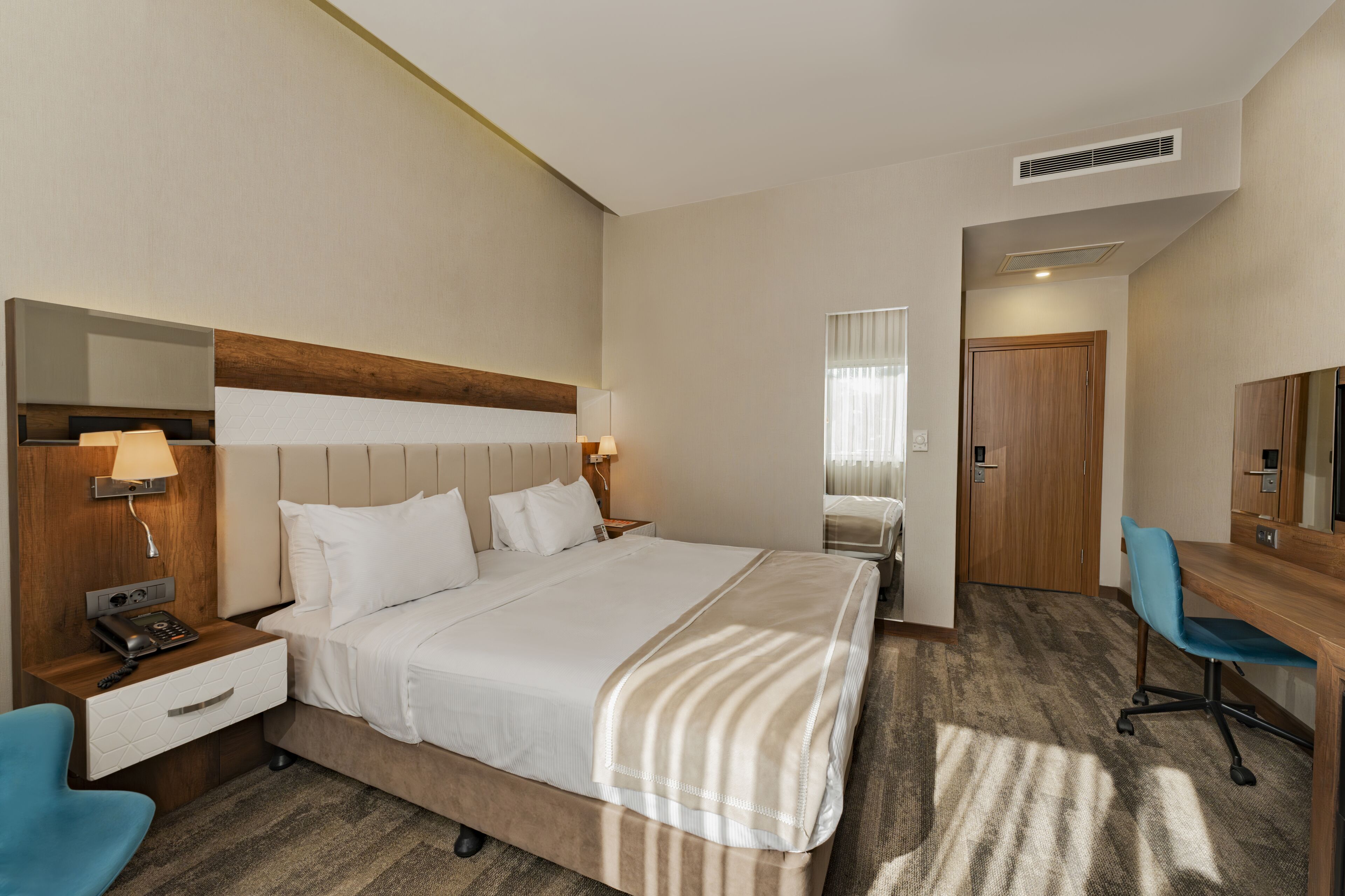 Habitación de hotel con una cama, mesita de noche con lámpara, escritorio con silla, espejo y puerta.