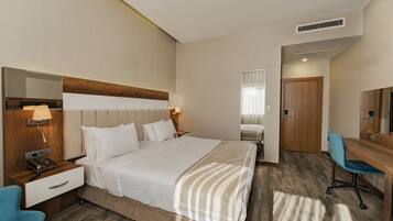 Habitación de hotel con una cama, mesita de noche con lámpara, escritorio con silla, espejo y puerta.
