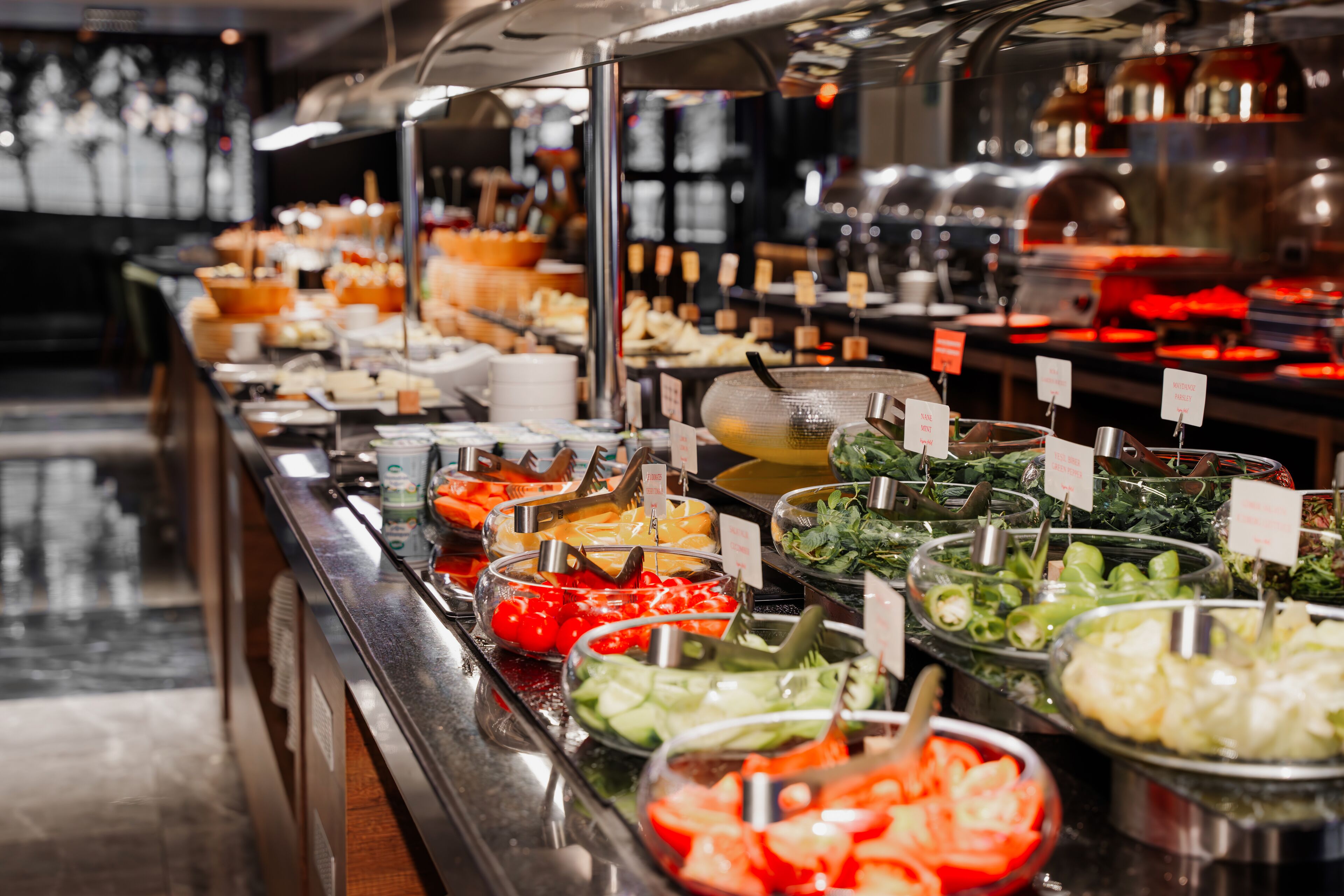 Un bufé con diversas opciones de comida, como ensaladas, verduras y carnes, dispuestas en recipientes de vidrio con precios a la vista.