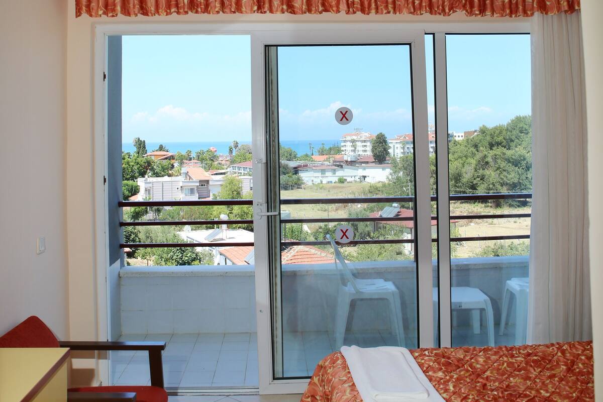 A hotel room with a balcony, a bed with a patterned bedspread, a desk with a chair, and a view of the surrounding area.