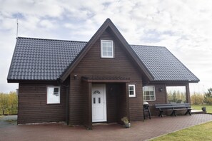 A brown wooden building with a tiled roof, a white door, and a small porch area with a bench and a table.