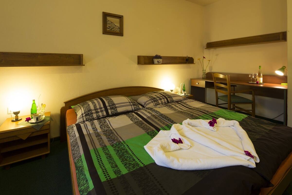 A neatly made bed with a patterned bedspread, a bedside table with a bottle and glasses, and a desk with a chair.