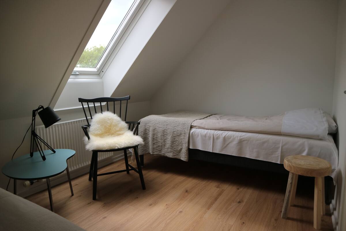 A small, well-lit room with a sloped ceiling, a single bed, a chair with a fluffy cushion, a small table, and a radiator.