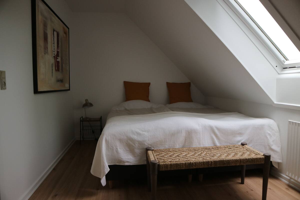 A bedroom with a sloped ceiling, a bed with white bedding, two brown pillows, a woven bench, and a painting on the wall.