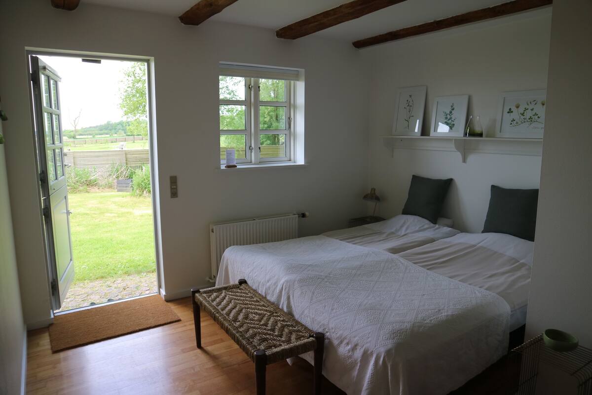 A bedroom with a bed, a bench, a window, and a door leading to a garden.