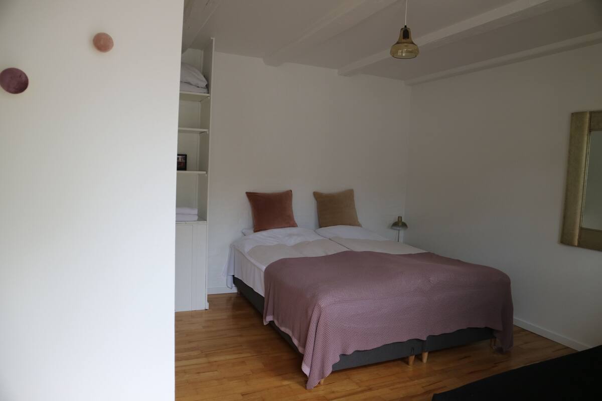 A bedroom with a bed, wooden flooring, a hanging light fixture, and a white wall.