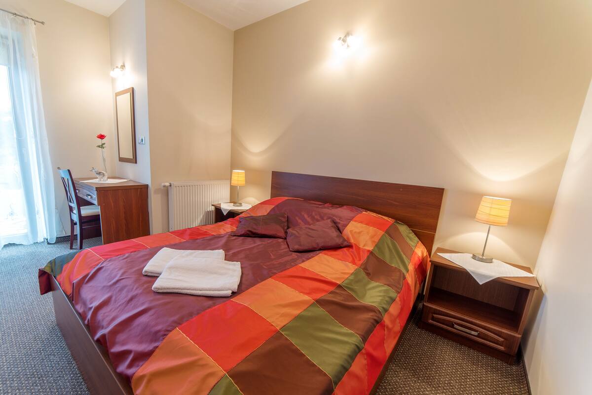 A neatly made bed with a colorful quilt, a bedside table with a lamp, and a chair in a hotel room.