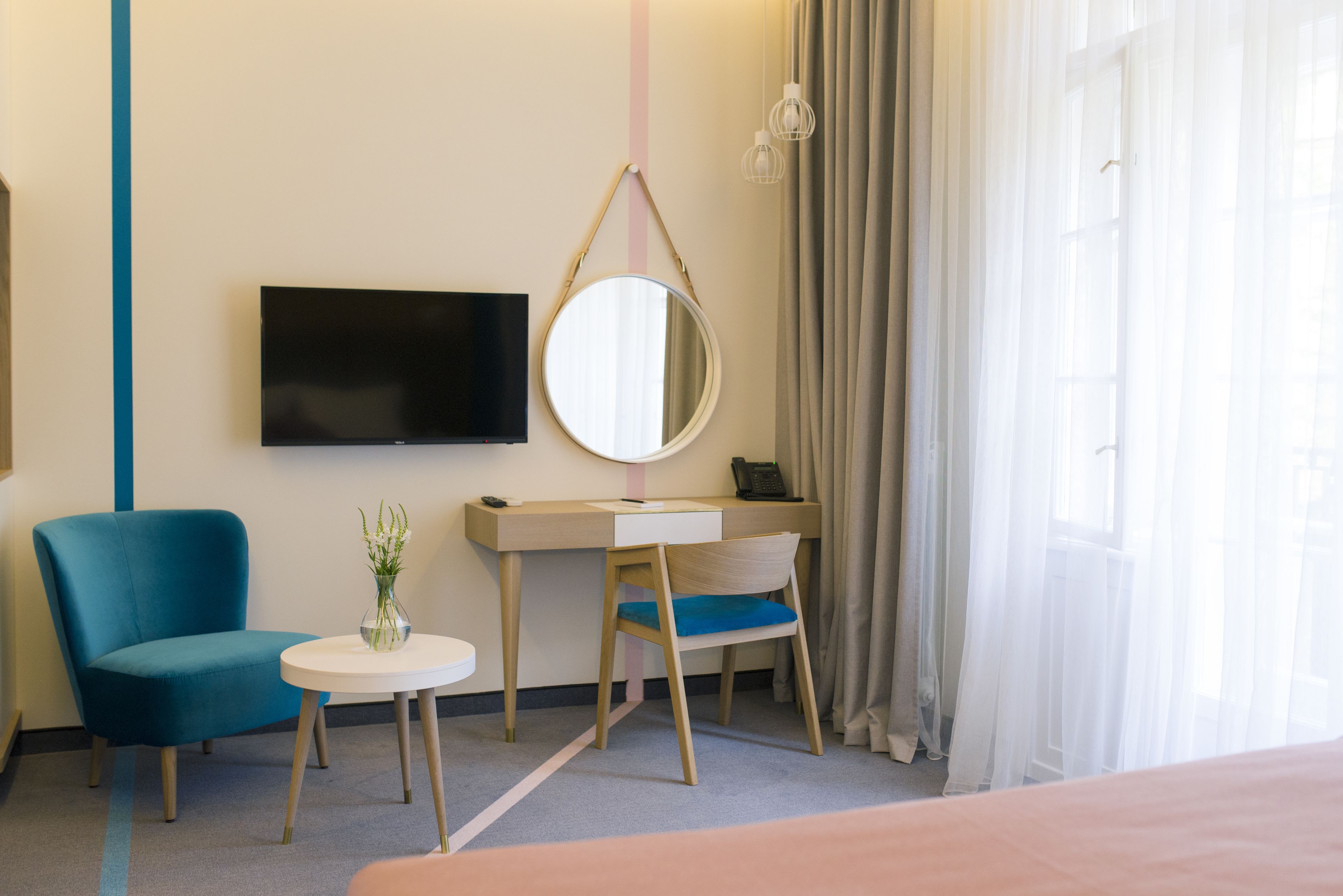Habitación de hotel con cama, escritorio con silla, televisión, espejo redondo, mesita con un florero y cortinas traslúcidas.