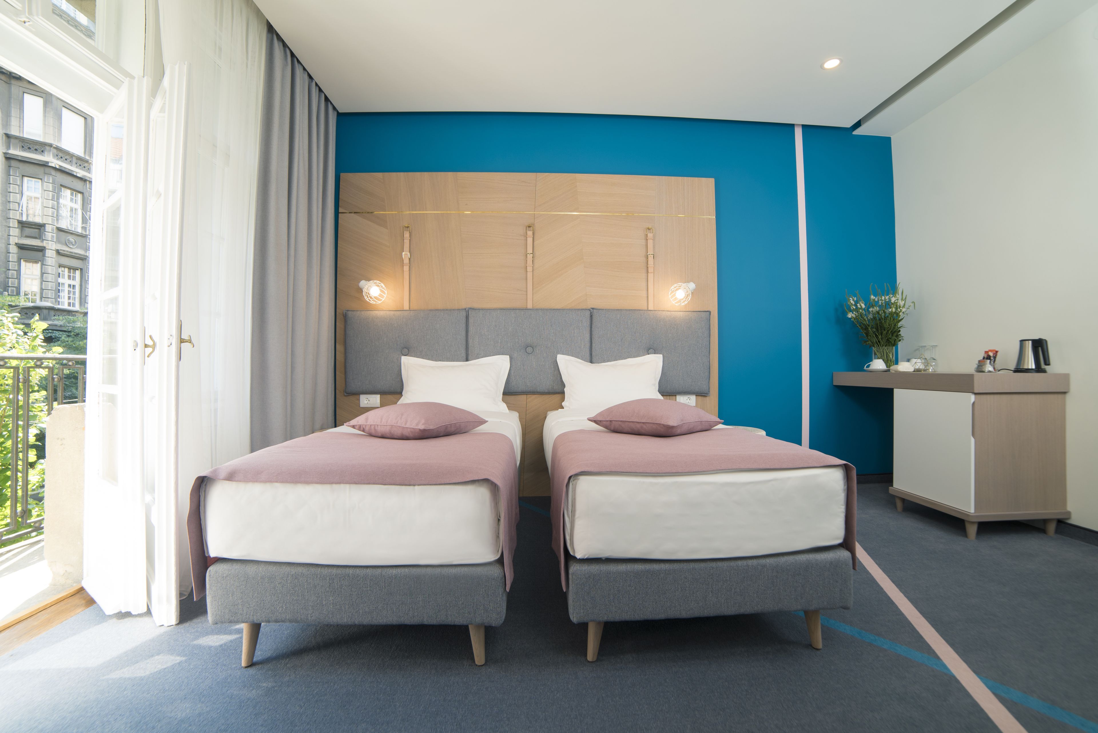A modern hotel room with two beds, a desk, and a view of the city.