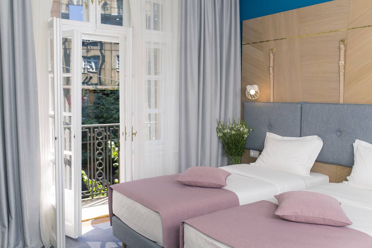 Habitación de hotel con balcón, cama con ropa de cama blanca y almohadas rosas, y cabecera gris.