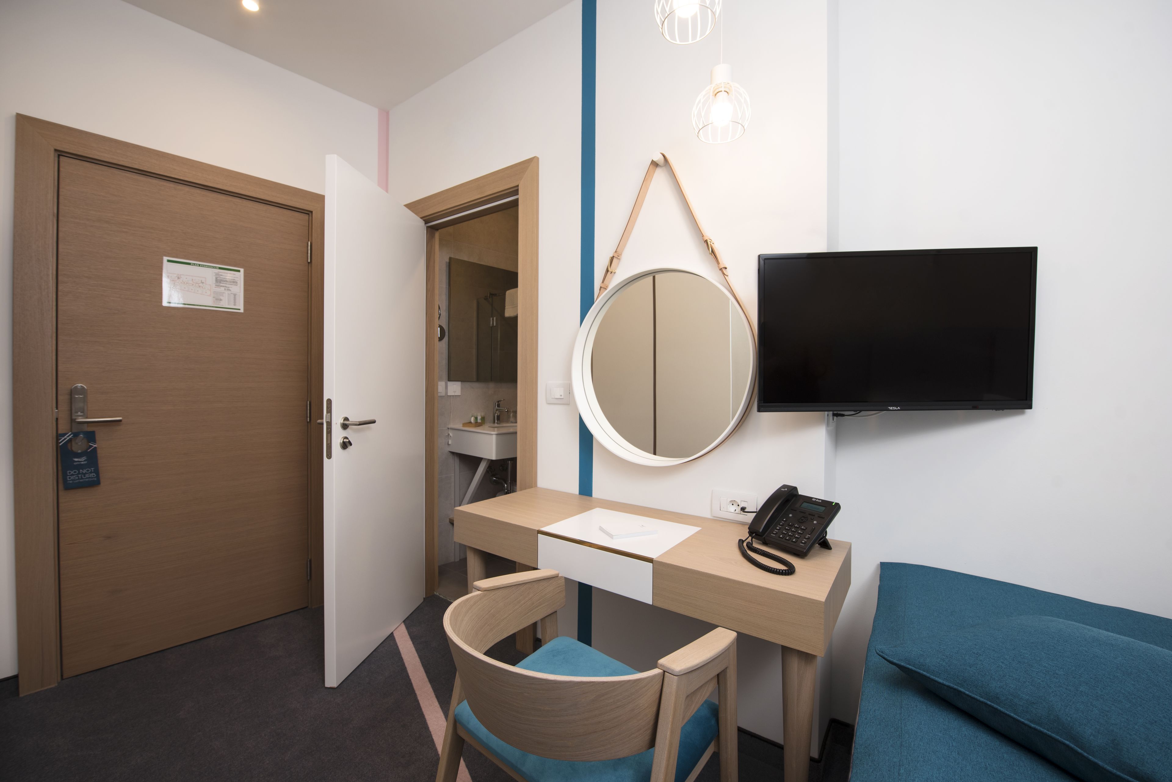 A hotel room with a wooden desk, a chair, a bed, a TV, a mirror, and a door.