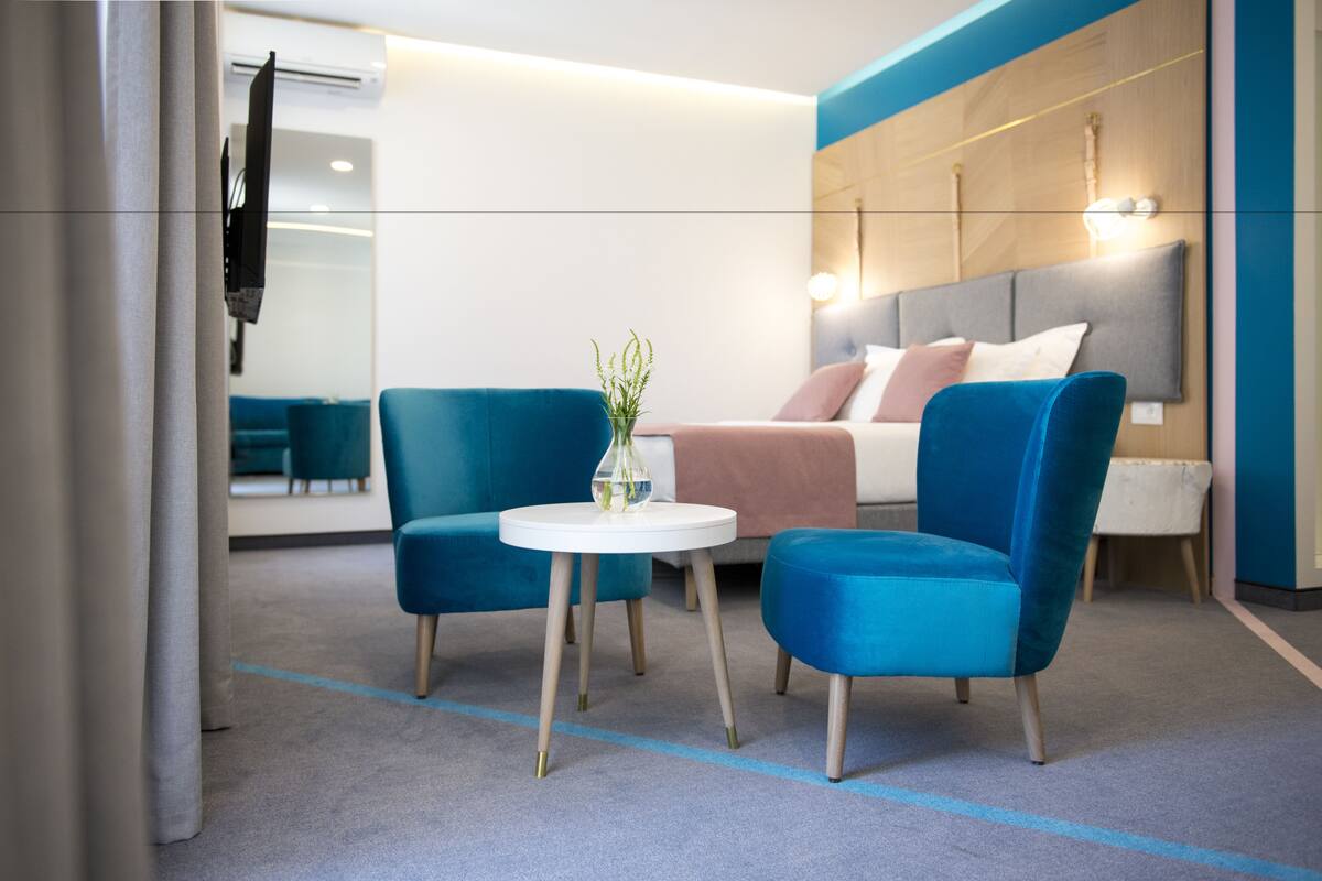 Una habitación de hotel moderna con una cama, dos sillones azules, una pequeña mesa blanca y un espejo.