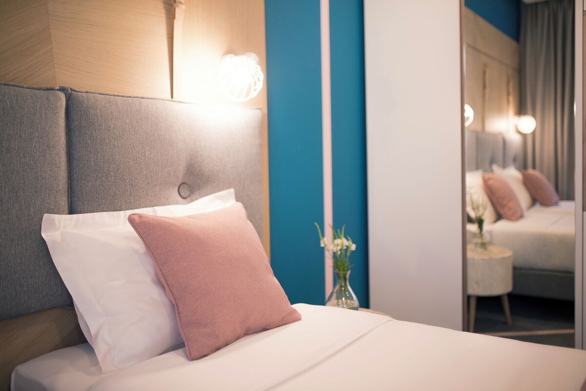 Ein ordentlich bezogenes Bett mit weißen und rosa Kissen, eine Blumenvase und ein Spiegel, der Bett und Vorhänge reflektiert.