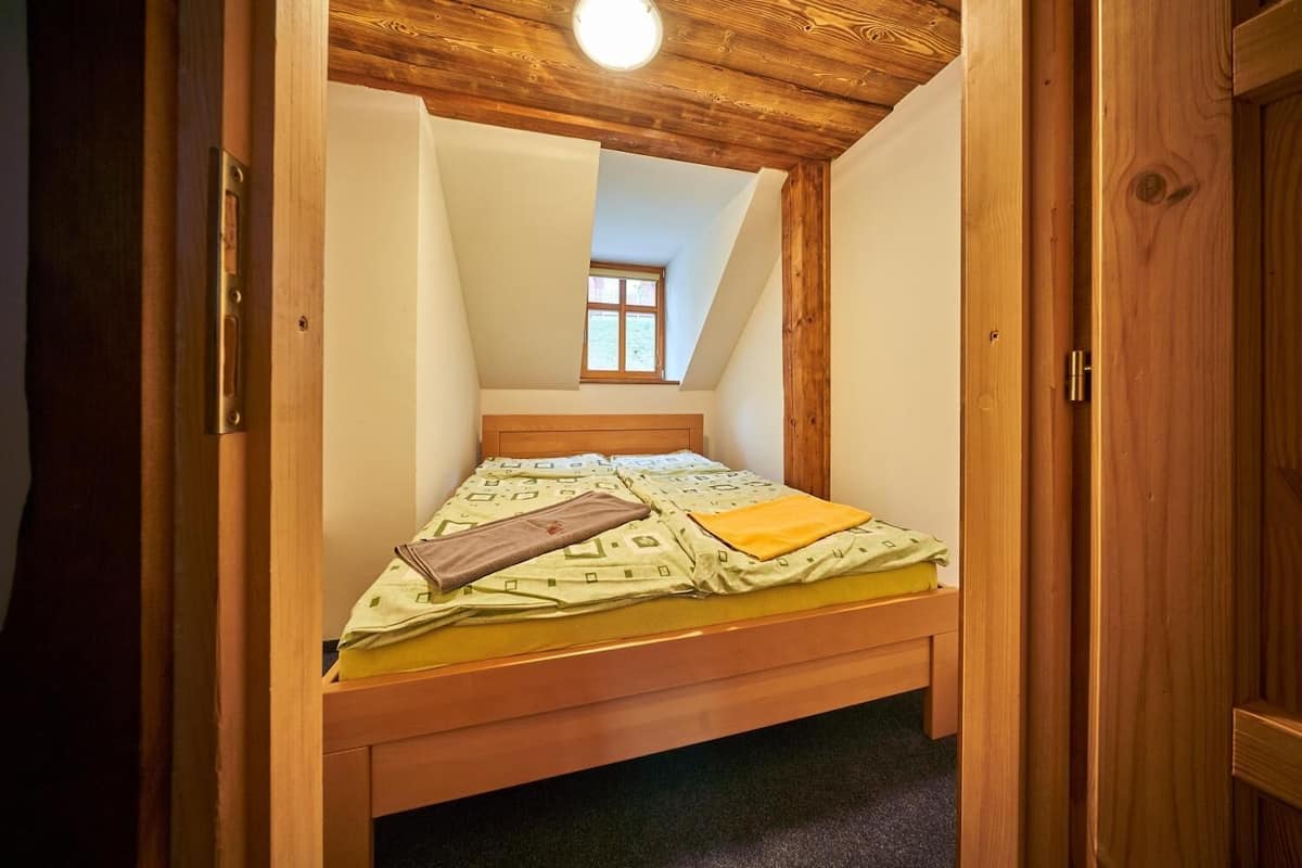 Ein schmales Zimmer mit schrägem Dach, einem Bett mit gemustertem Überwurf und einem Fenster am Ende.