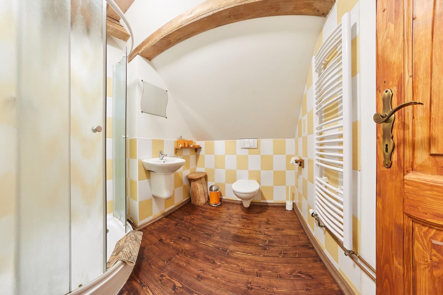 A narrow bathroom with a wooden floor, a sink, a toilet, and a wooden door with a brass handle.