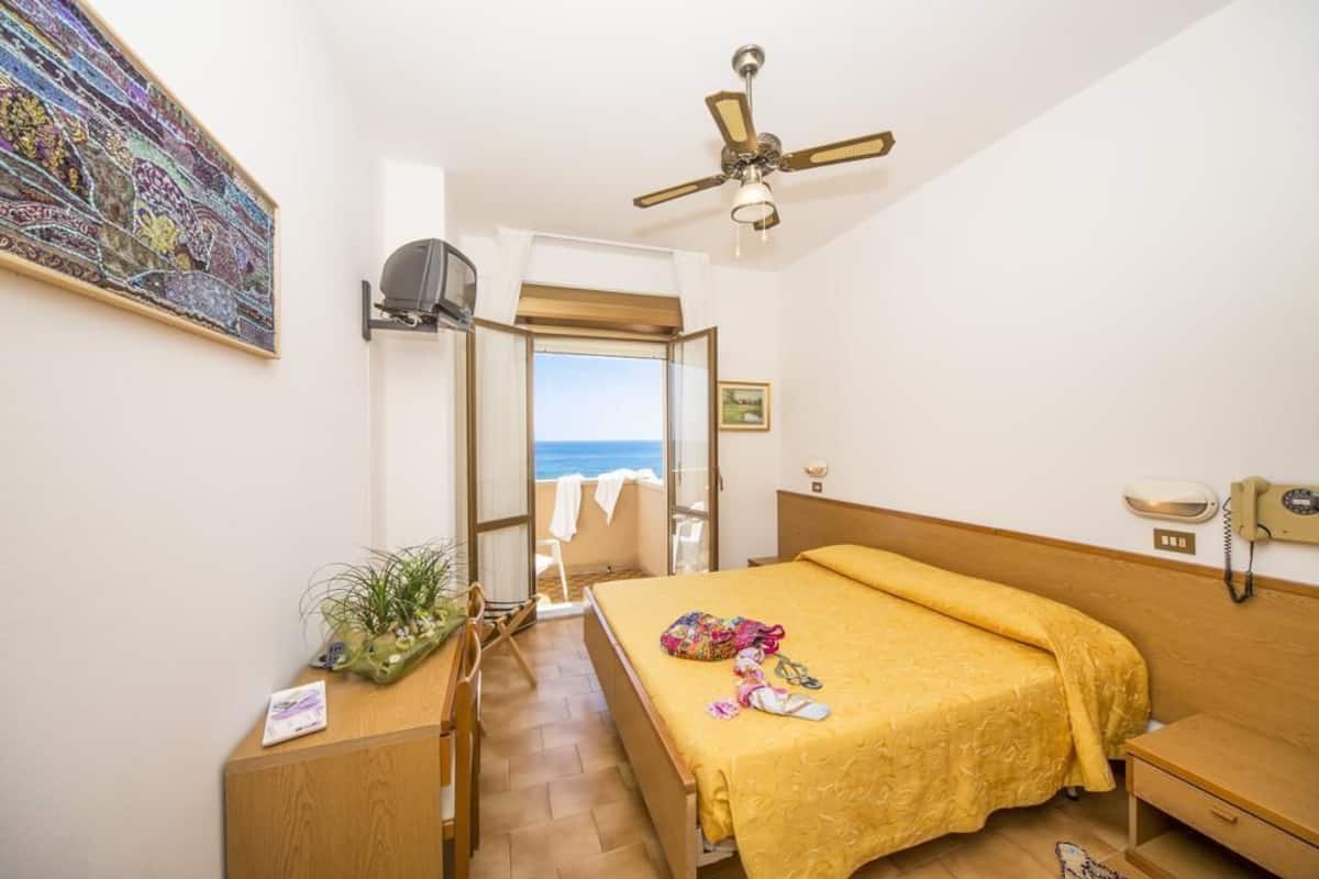 Une chambre à coucher avec un lit, un ventilateur de plafond, un téléphone sur la table de chevet, une fenêtre donnant sur la mer et un tableau accroché au mur.