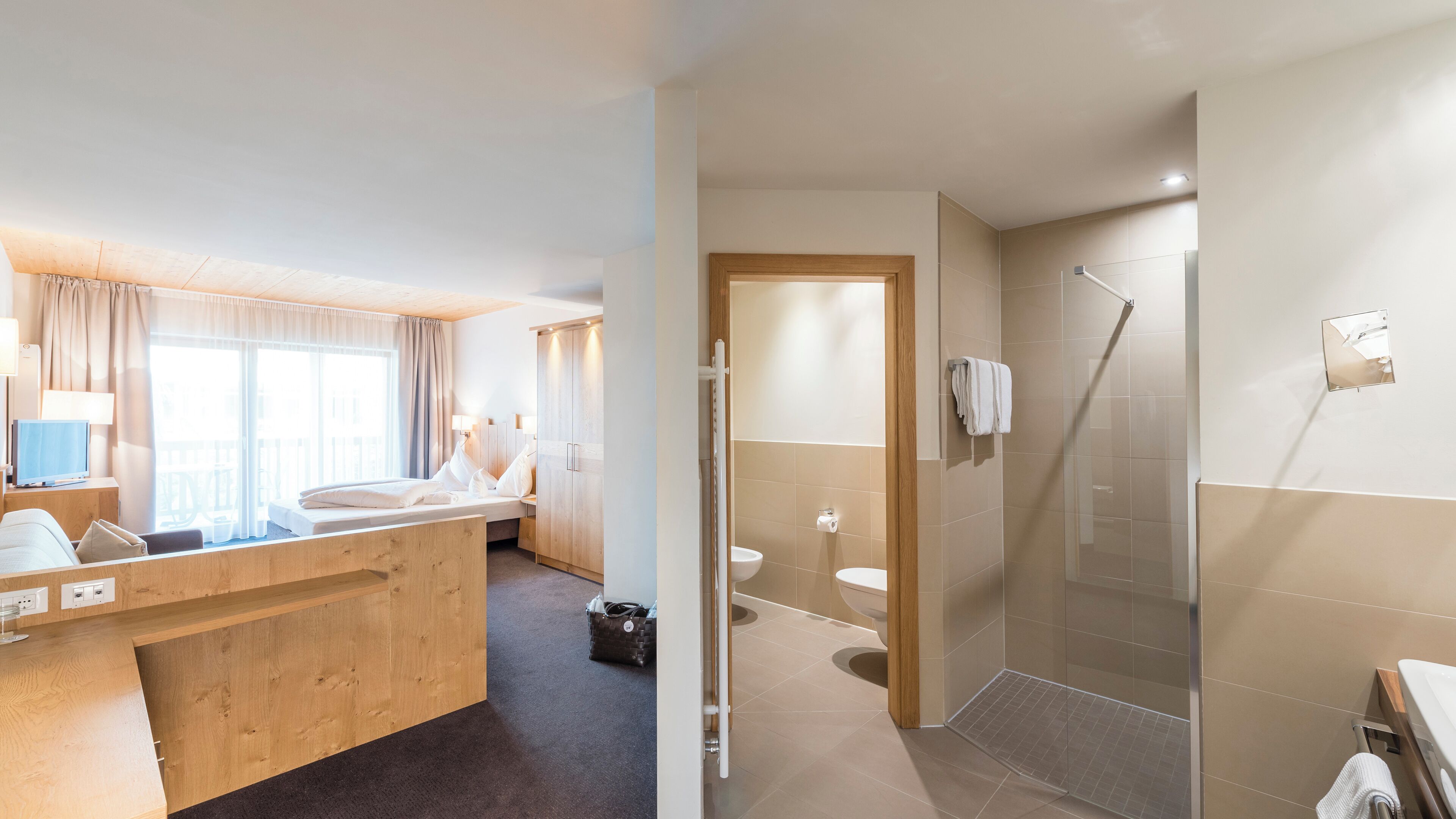 A modern hotel room with a wooden desk, a bed, a bathroom with a shower, and a toilet.