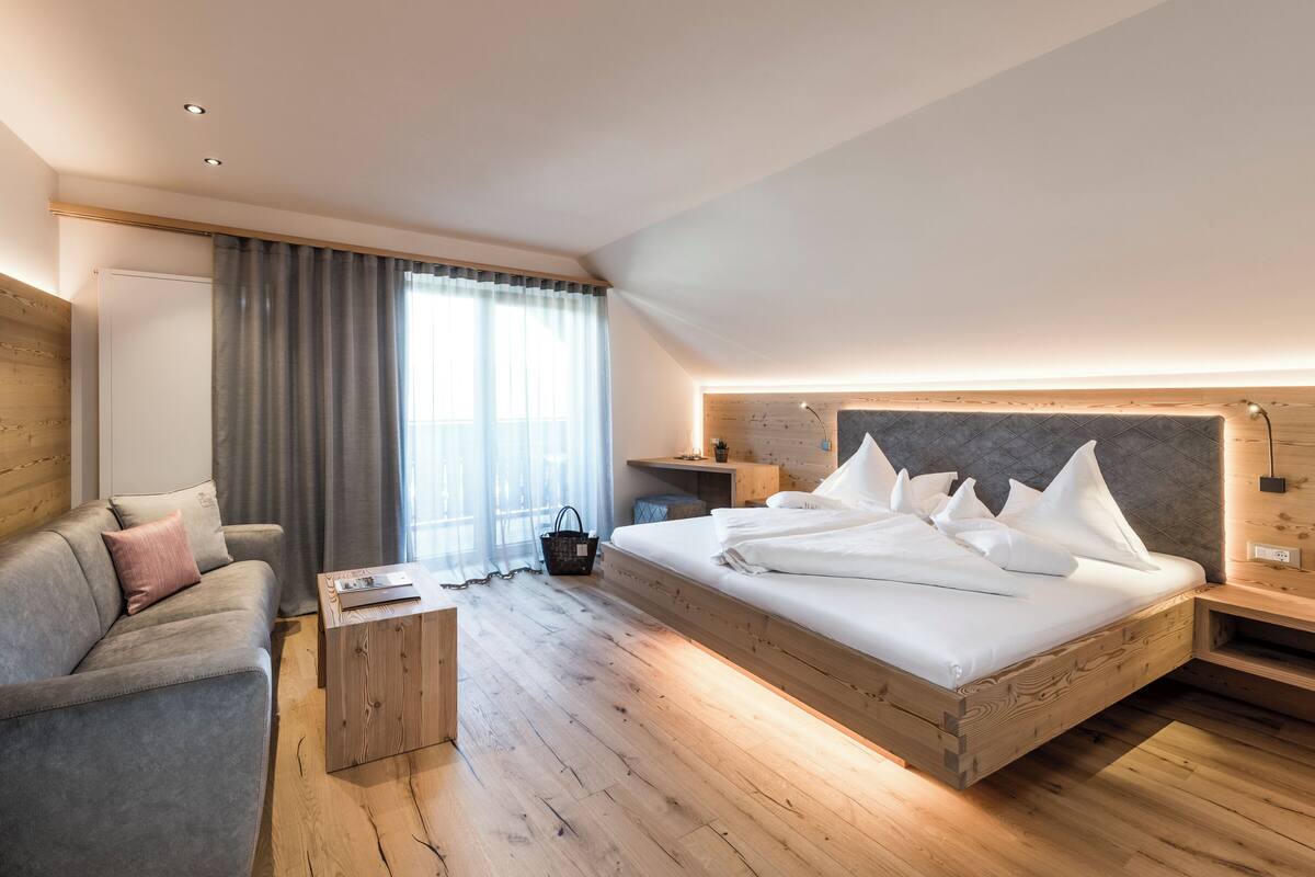 Een moderne hotelkamer met een houten vloer, een groot bed met witte lakens, een grijze bank en een klein houten bijzettafeltje.