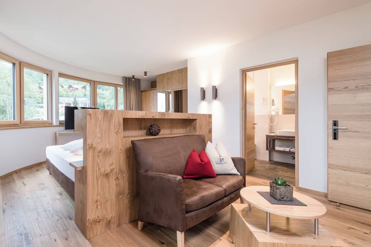 Een moderne kamer met een houten bed, een bruine leren bank met rode kussens, een klein rond tafeltje en een groot raam met uitzicht op bomen en gebouwen.