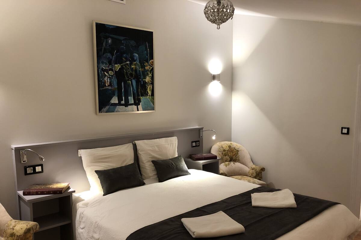 Un lit bien fait, recouvert d’une couvre-lit noir et blanc, une petite table de chevet avec un livre, un fauteuil décoratif et un tableau accroché au mur.