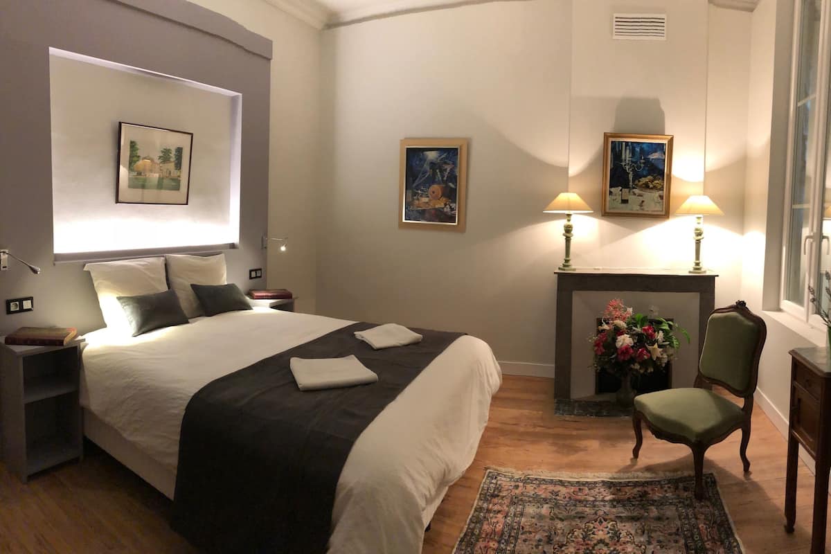 Une chambre d’hôtel avec un grand lit, des lampes de chevet, une cheminée, un fauteuil et une petite table sur laquelle est posée une plante.