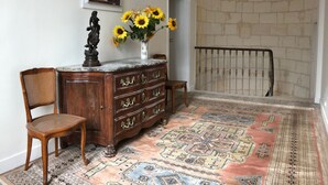 Een kamer met een houten dressoir, een stoel, een vaas met zonnebloemen, een kamerbreed tapijt, een schilderij aan de muur en een stenen muur.
