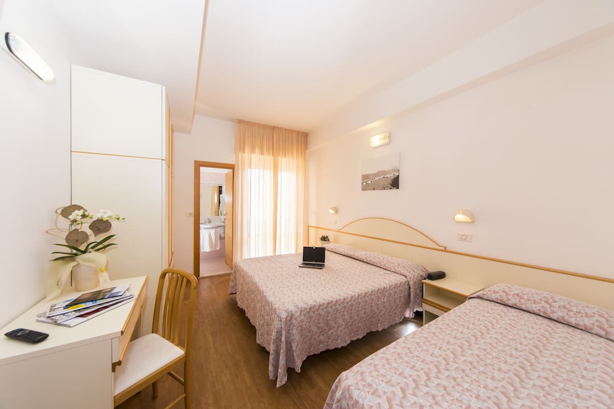 Une chambre d’hôtel équipée d’un bureau, de deux lits, d’une chaise, d’un ordinateur portable et d’une petite table sur laquelle est posé un vase de fleurs.