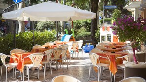 Een terras met tafels en stoelen, een witte parasol en een blauw bord met richtingaanwijzingen.