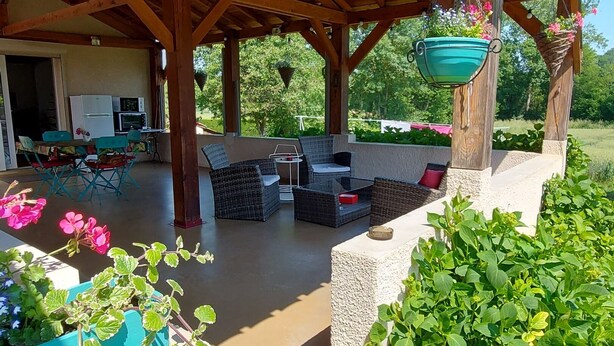 Ein überdachter Außenbereich mit Holzbalken, einer hängenden Pflanzbox mit Blumen und Blick auf ein Haus mit Gartenmöbeln.