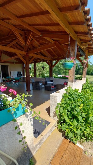 A covered outdoor seating area with wooden beams, a hanging planter with flowers, and a view of a house with outdoor furniture. - Le Moulin de L'étang (Chaneins)