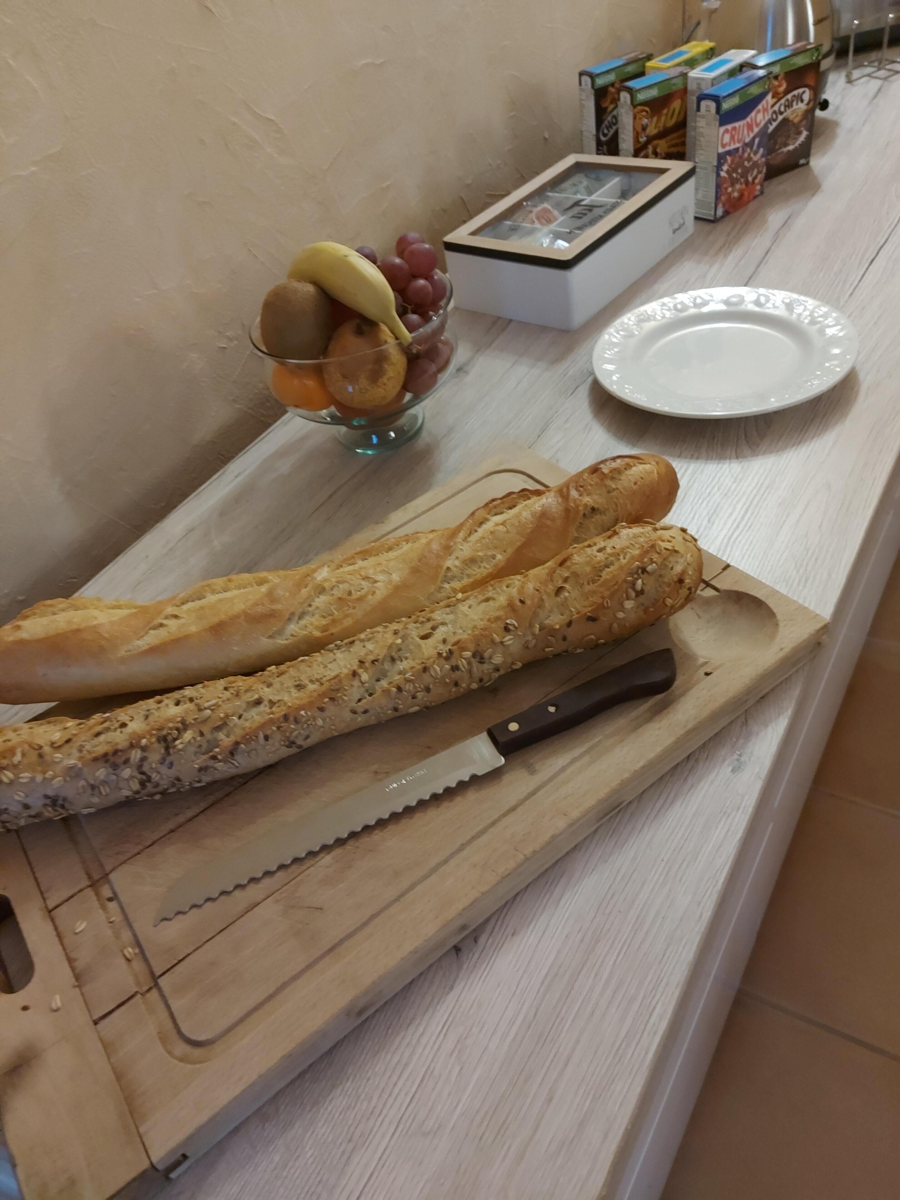 En træskærebræt med et baguette og en kniv, en glasskål med frugt og cornflakes på en køkkenbordplade.