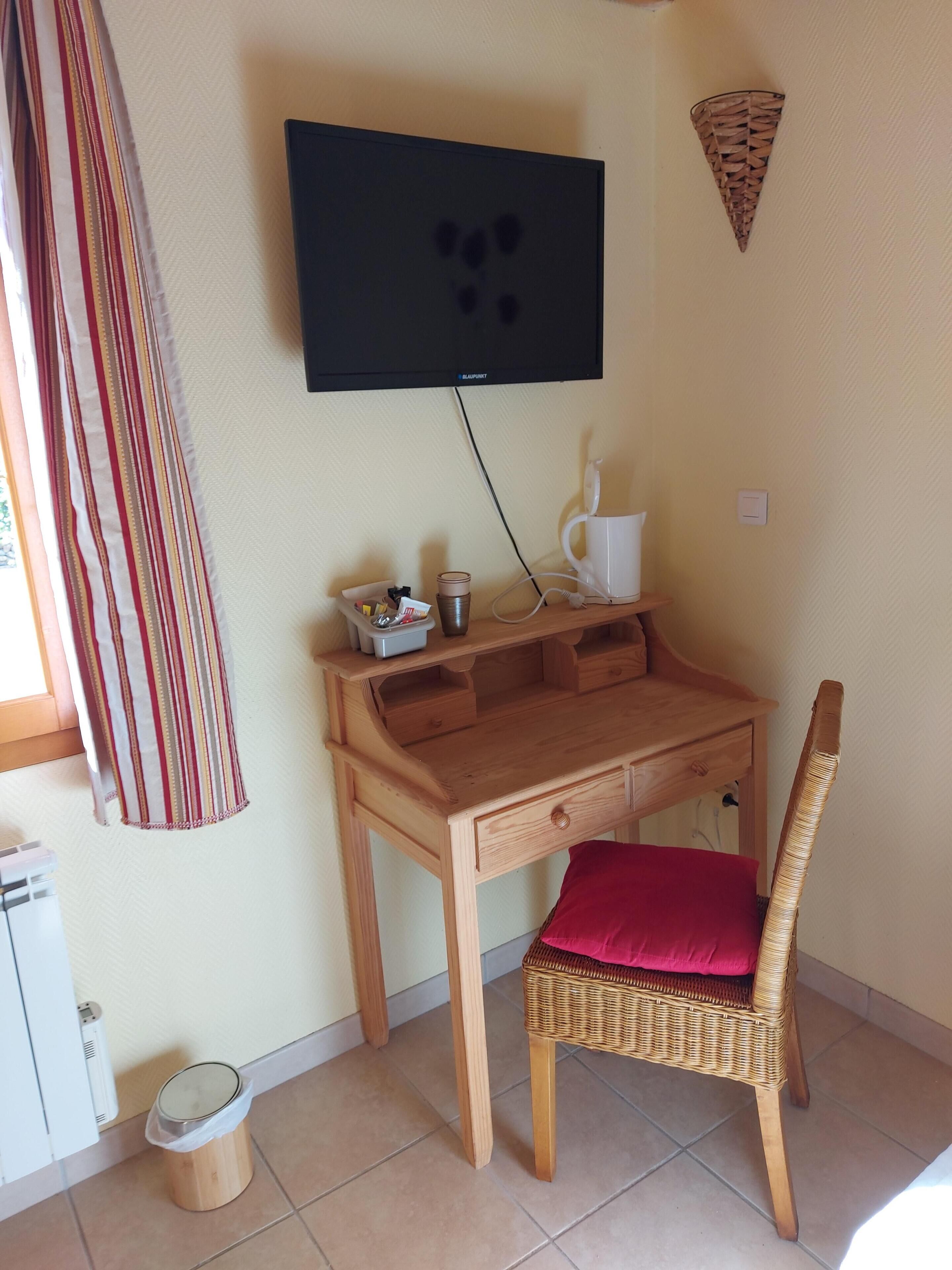 A small room with a wooden desk, a wicker chair with a red cushion, a television mounted on the wall, a small trash bin, and a window with a striped curtain.