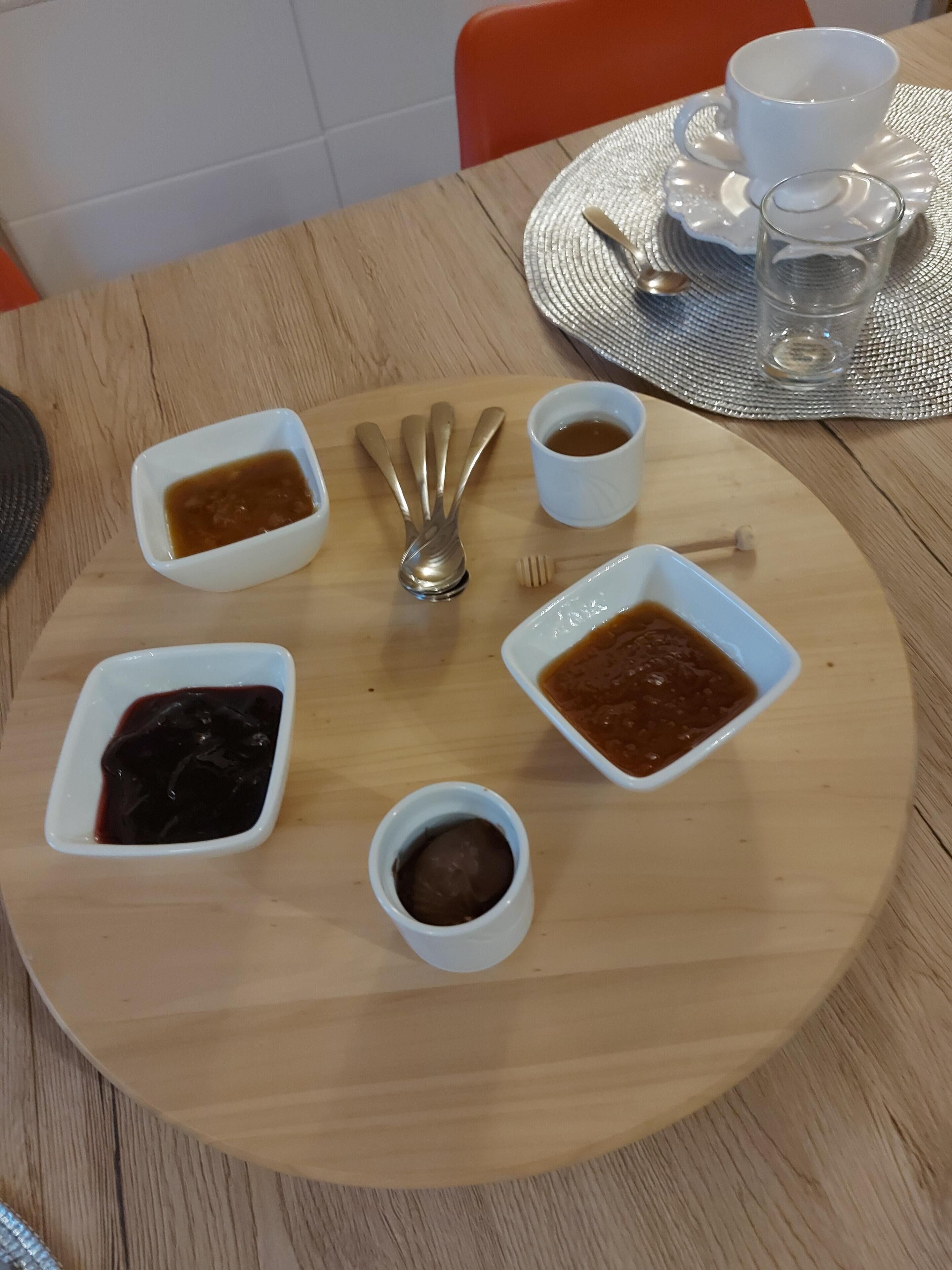 Una mesa de madera con un mantel individual blanco, seis recipientes pequeños blancos con diferentes salsas, una cuchara y una taza y platillo blancos sobre una bandeja de plata.