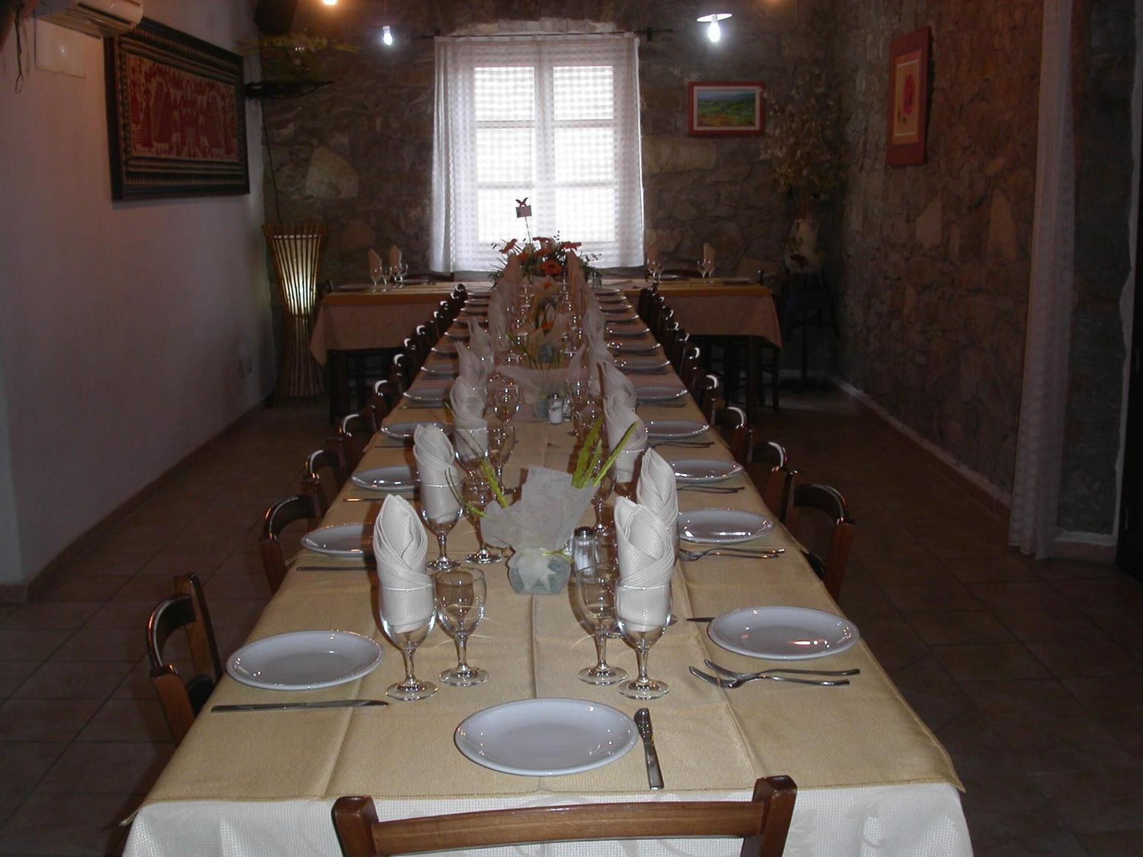 A long dining table set for a meal with chairs on both sides, a chandelier, and a stone wall.