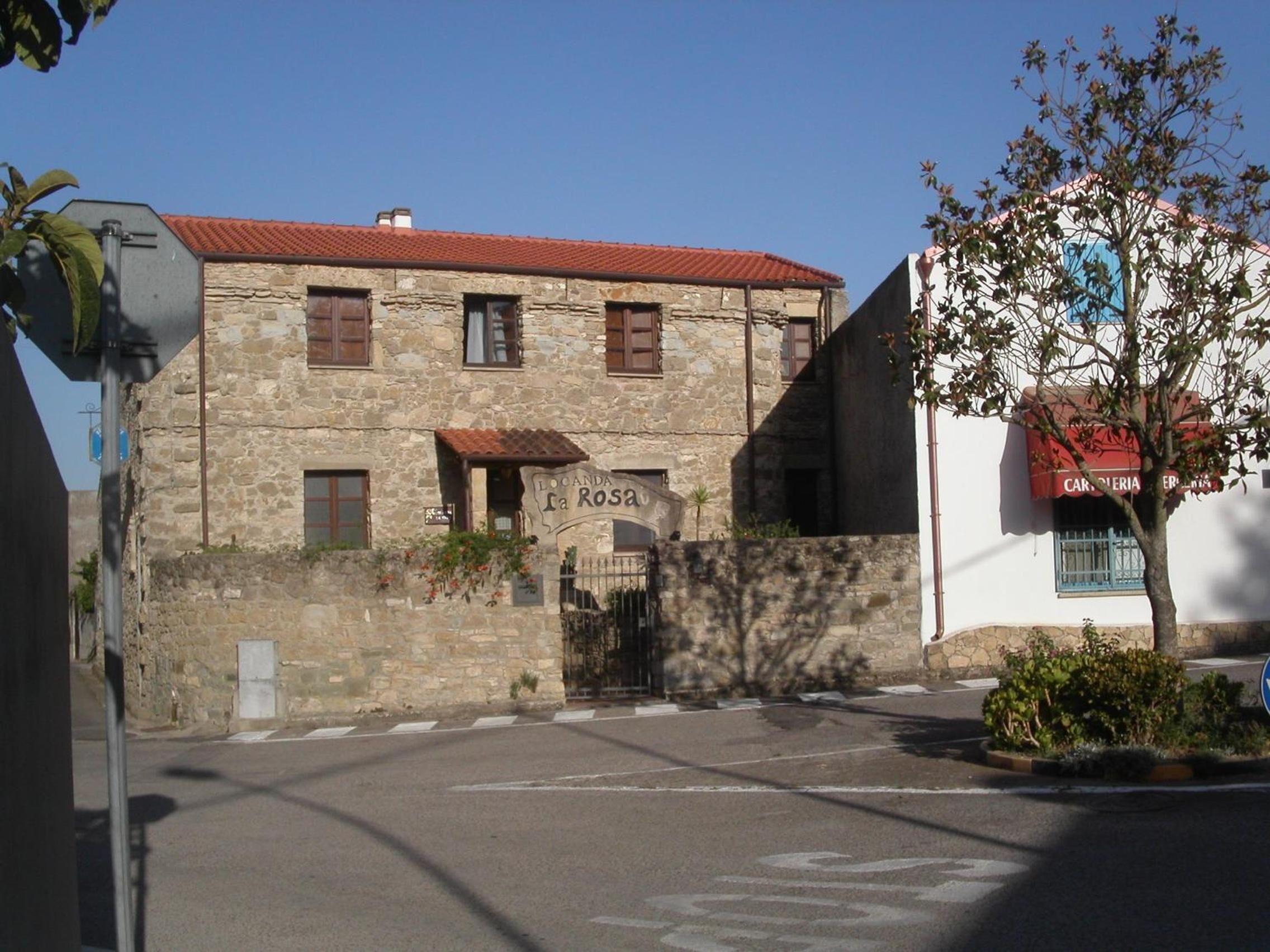 Un edificio in pietra con tetto in tegole rosse, un cartello con su scritto “LOCA DE LA ROSA” e un piccolo giardino con un albero.