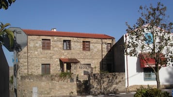 Un edificio in pietra con tetto in tegole rosse, un cartello con su scritto “LOCA DE LA ROSA” e un piccolo giardino con un albero.