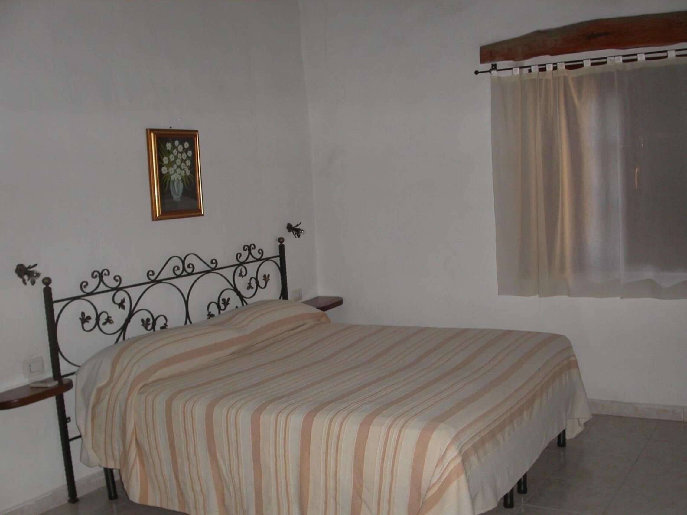 A bedroom with a bed, a bedside table, a nightstand, a window with a curtain, and a framed picture on the wall.