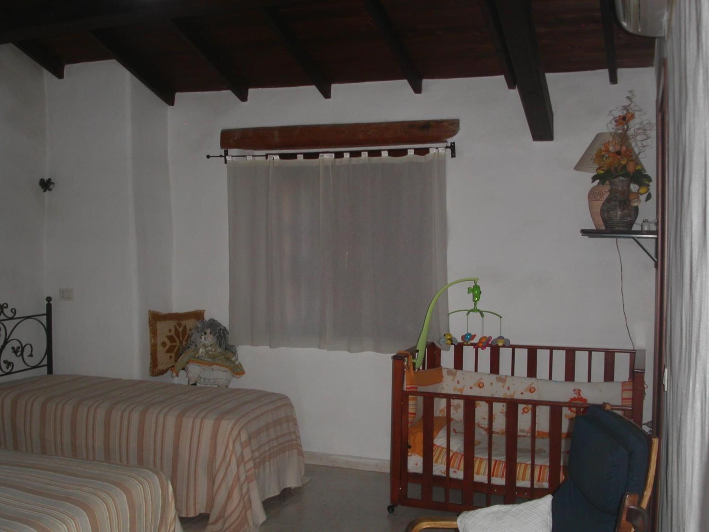 A bedroom with two beds, a crib, a window with curtains, a wall-mounted shelf with a vase, and a wooden ceiling.