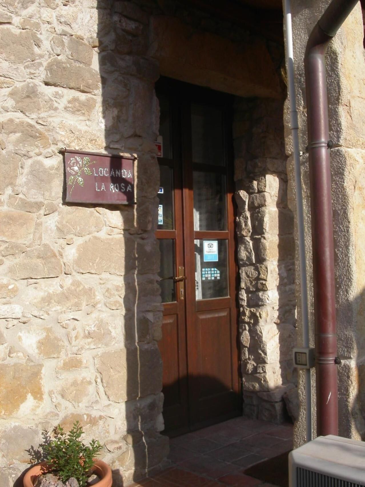 Entrada de un edificio de piedra con una puerta de madera, un letrero que dice "LOCANDA LA ROSA" y una planta en maceta.