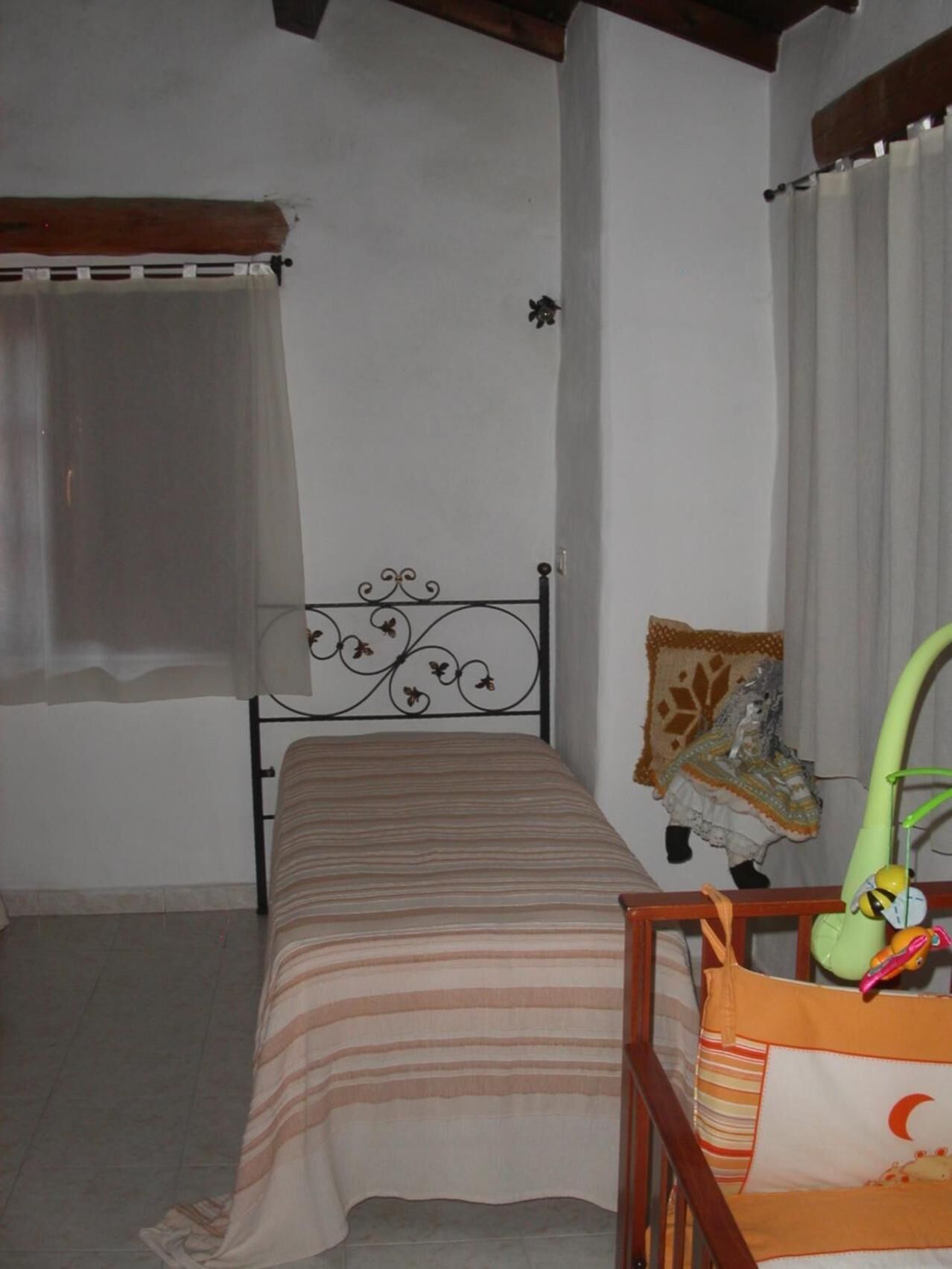 A small, single-bed room with a metal headboard, a bedside table with a lamp, and a window with sheer curtains.