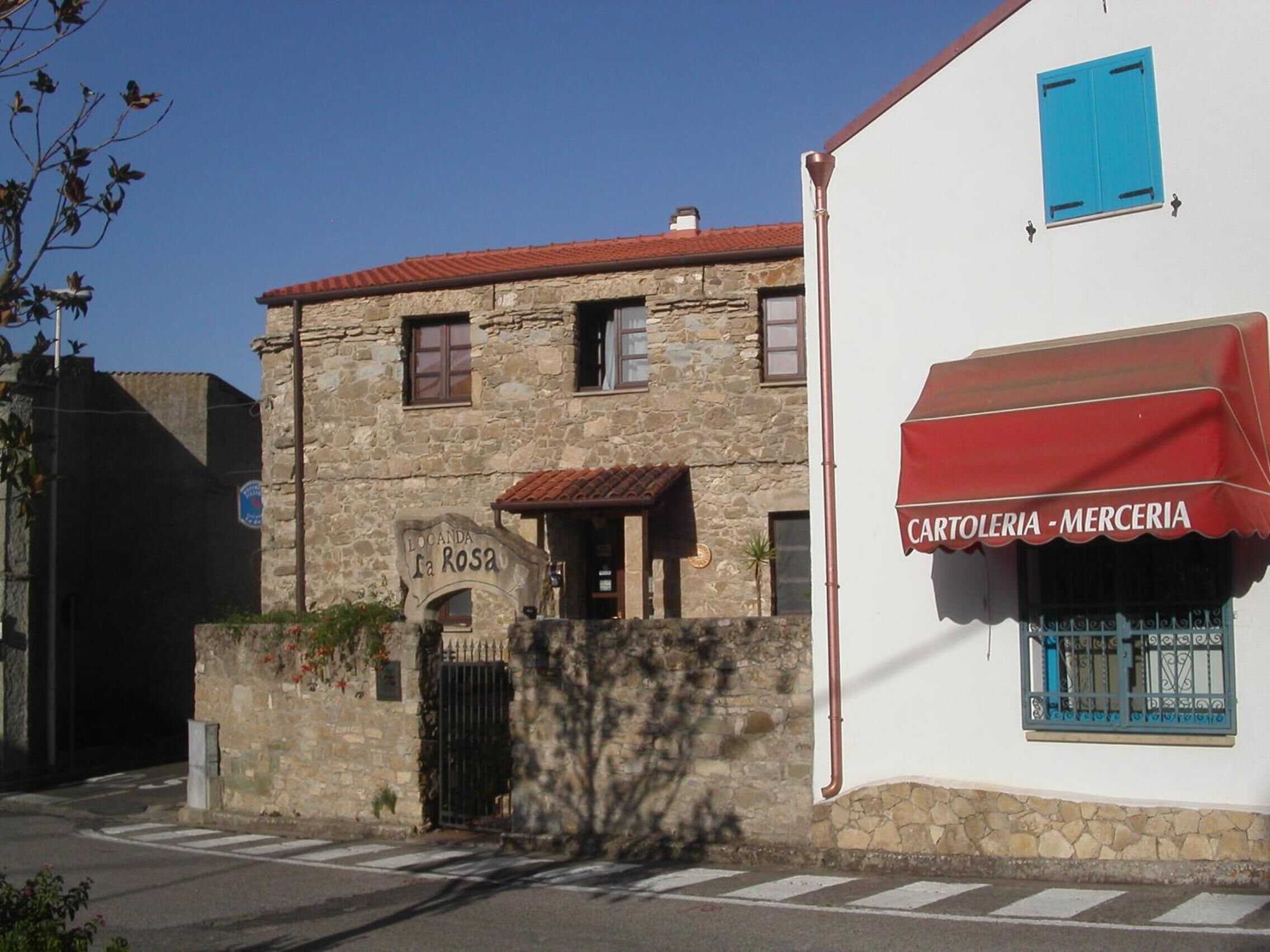Un edificio de piedra con un letrero que dice "Locanda La Rosa" y un toldo rojo con la inscripción "Cartoleria -Merceria".